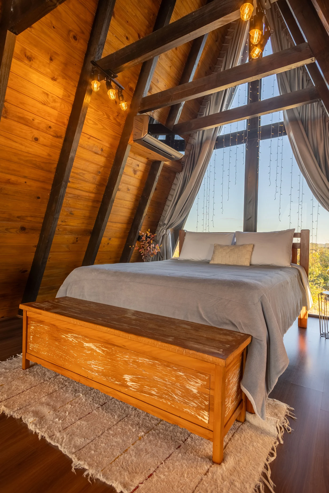 Double bed, wooden chest and cream rug beneath a tall triangular curtained window
